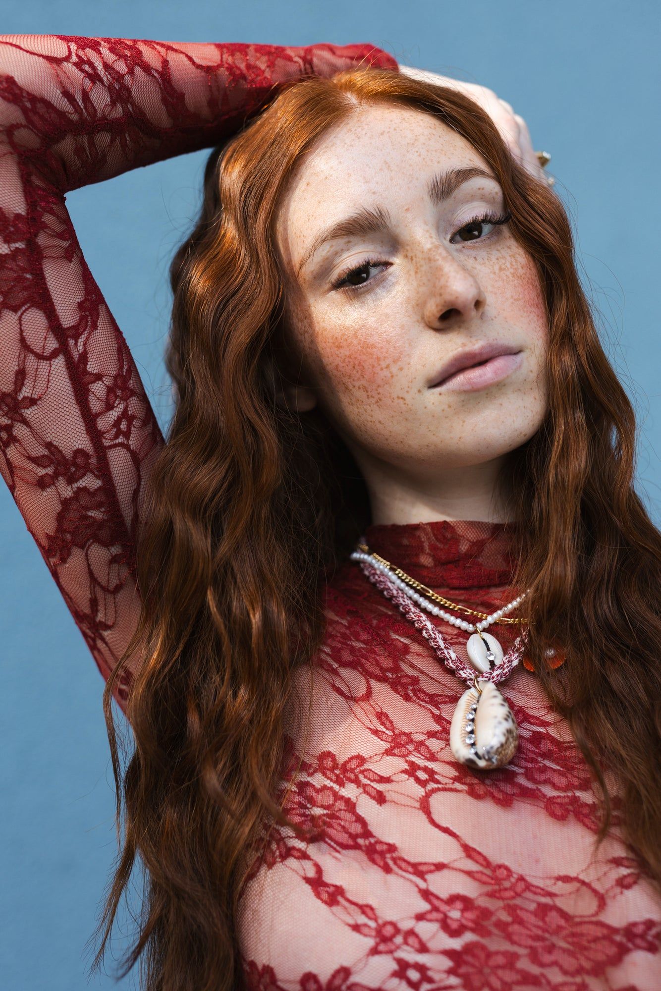 A person with long red hair wearing a red lace top and the WALD Berlin La Piscina Bootylicious Necklace With Ribbon poses against a blue background with one arm raised.