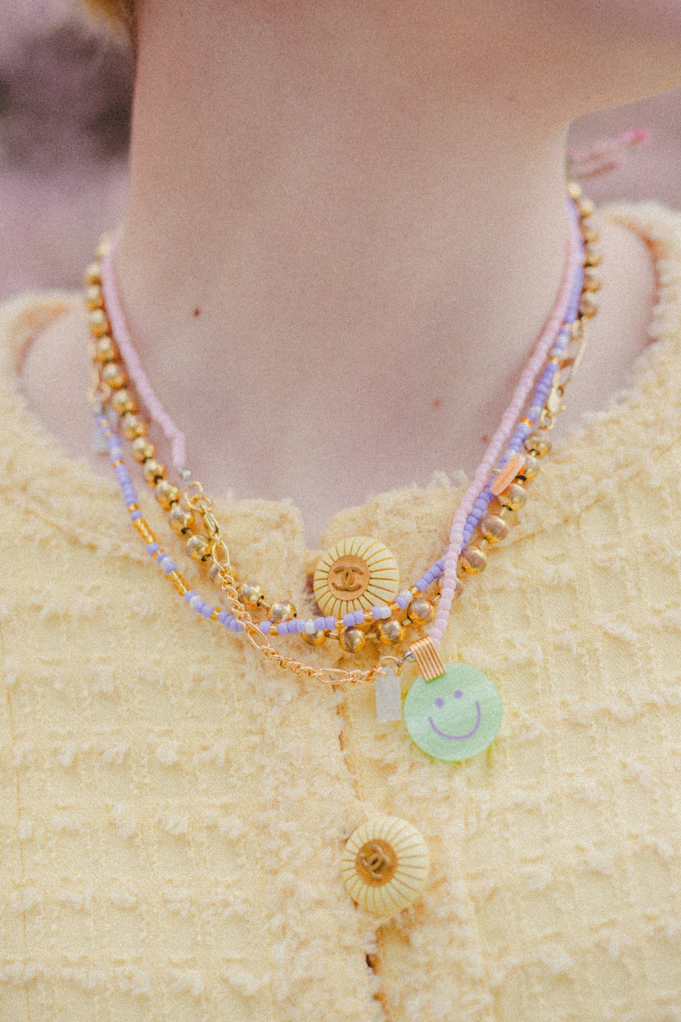 A person wears a yellow textured garment with layered necklaces, including beaded 18k gold plated pieces and WALD Berlin's Pop Smilie Mini necklace featuring a green Smilie Medaillon pendant.