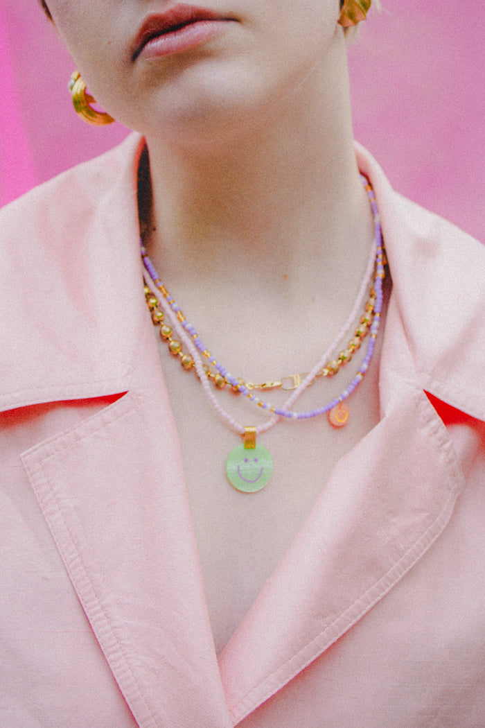 Person wearing a pale pink collared jacket and layered colorful necklaces, including the WALD Berlin Pop Smilie Mini 18k gold plated medallion, against a pink background.