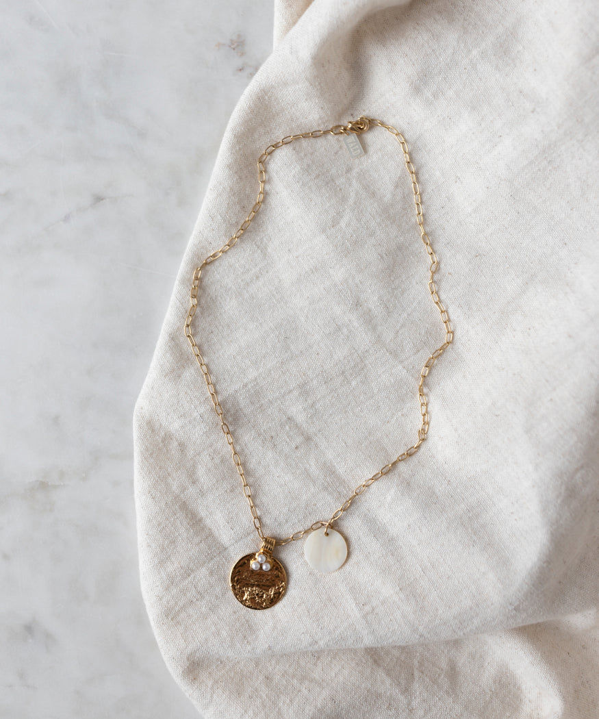 The Happy Holidays Medaillon Pearl Necklace by WALD Berlin is a delicate piece crafted from 18k gold-plated brass, featuring two elegant pendants—a textured gold disc and a smooth white disc with hints of sweetwater pearls—resting gracefully on a soft white fabric.