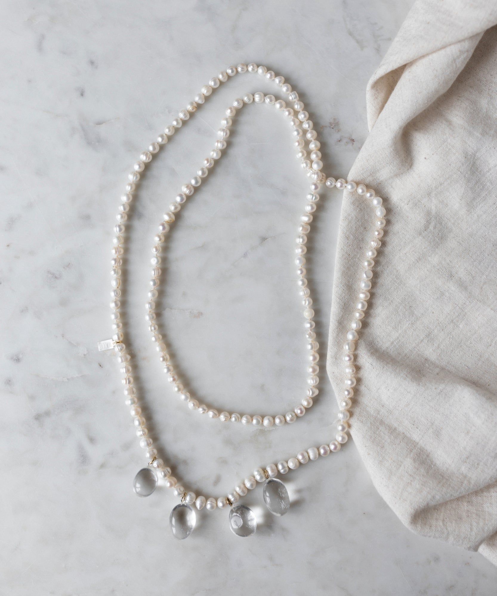 The Happy Holidays Long Pearl Drop Necklace by WALD Berlin, adorned with sweetwater pearls and three glass pendants, is gracefully displayed on a marble surface beside a piece of beige fabric.