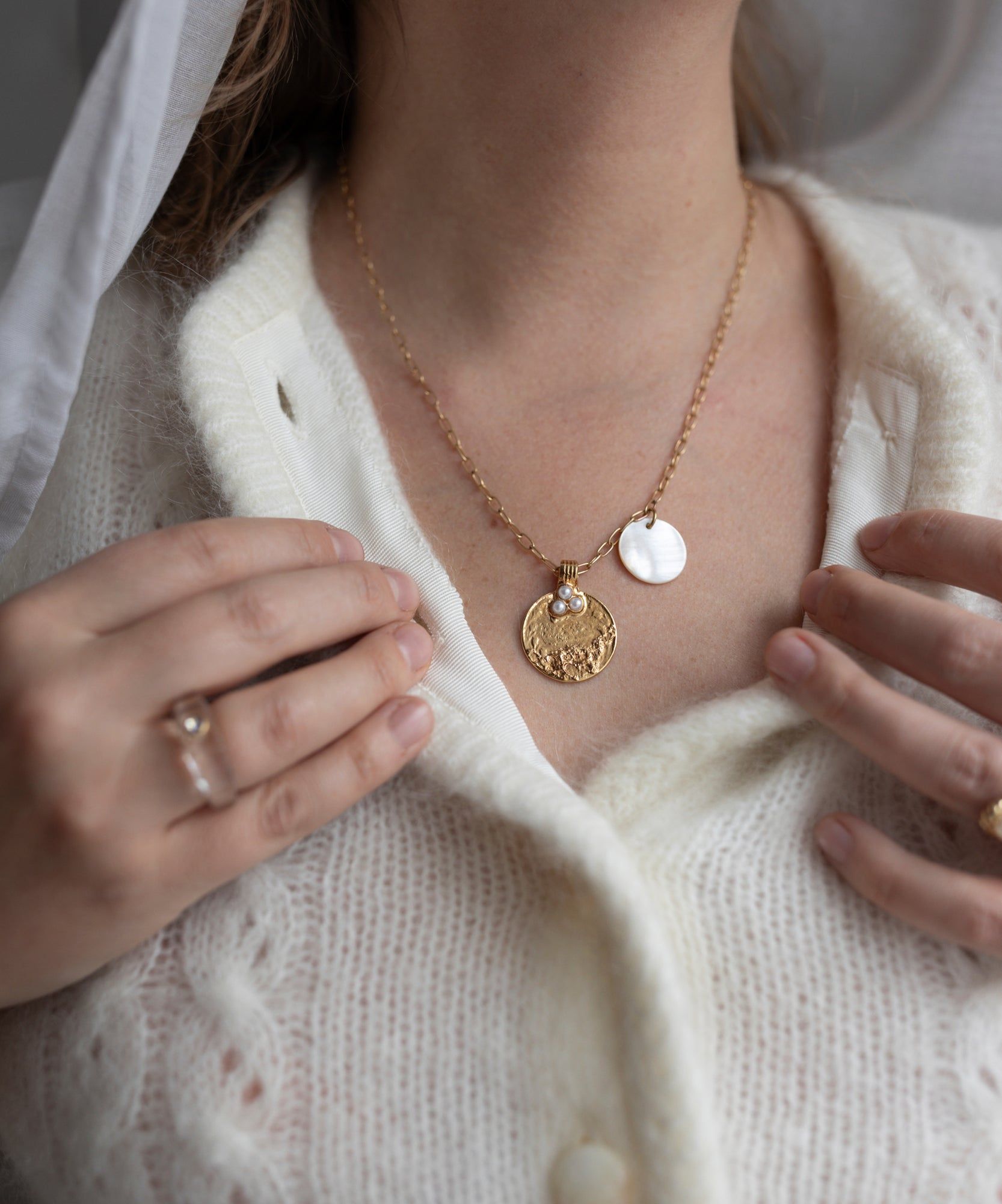 A person wearing a white knit sweater and the Happy Holidays Medaillon Pearl Necklace by WALD Berlin, adjusting the collar with hands adorned in rings of 18k gold-plated brass.
