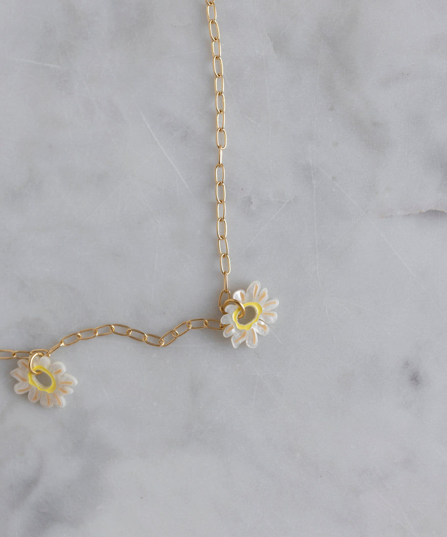 An 18k gold-plated chain necklace with two small daisy charms, each featuring white petals and a yellow center, is displayed on a light gray marble surface. This Daisy How High Necklace by WALD Berlin is perfect as an everyday accessory.