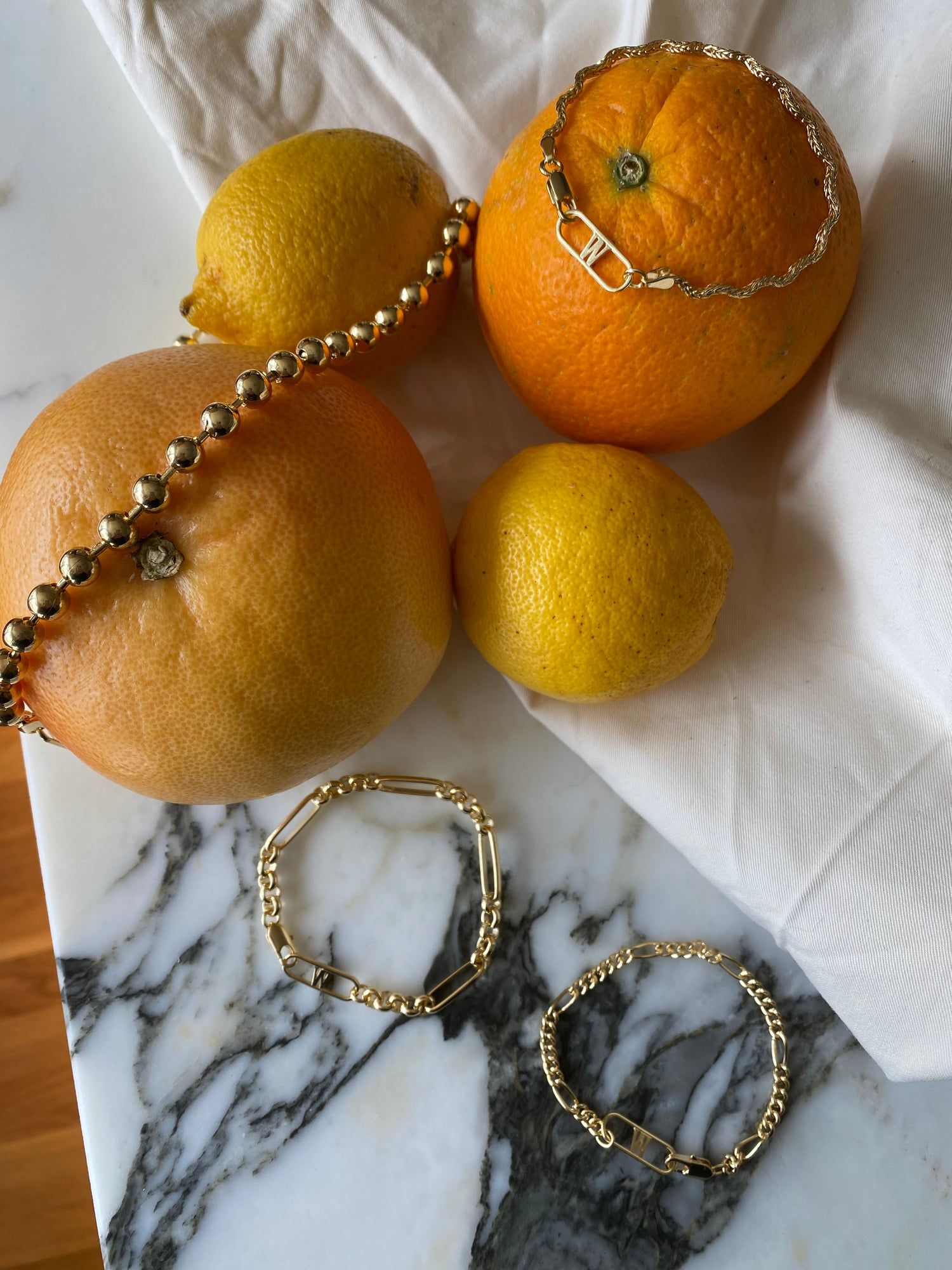 Irina Necklace Gold by WALD Berlin and brass bracelets alongside oranges on a marble table.