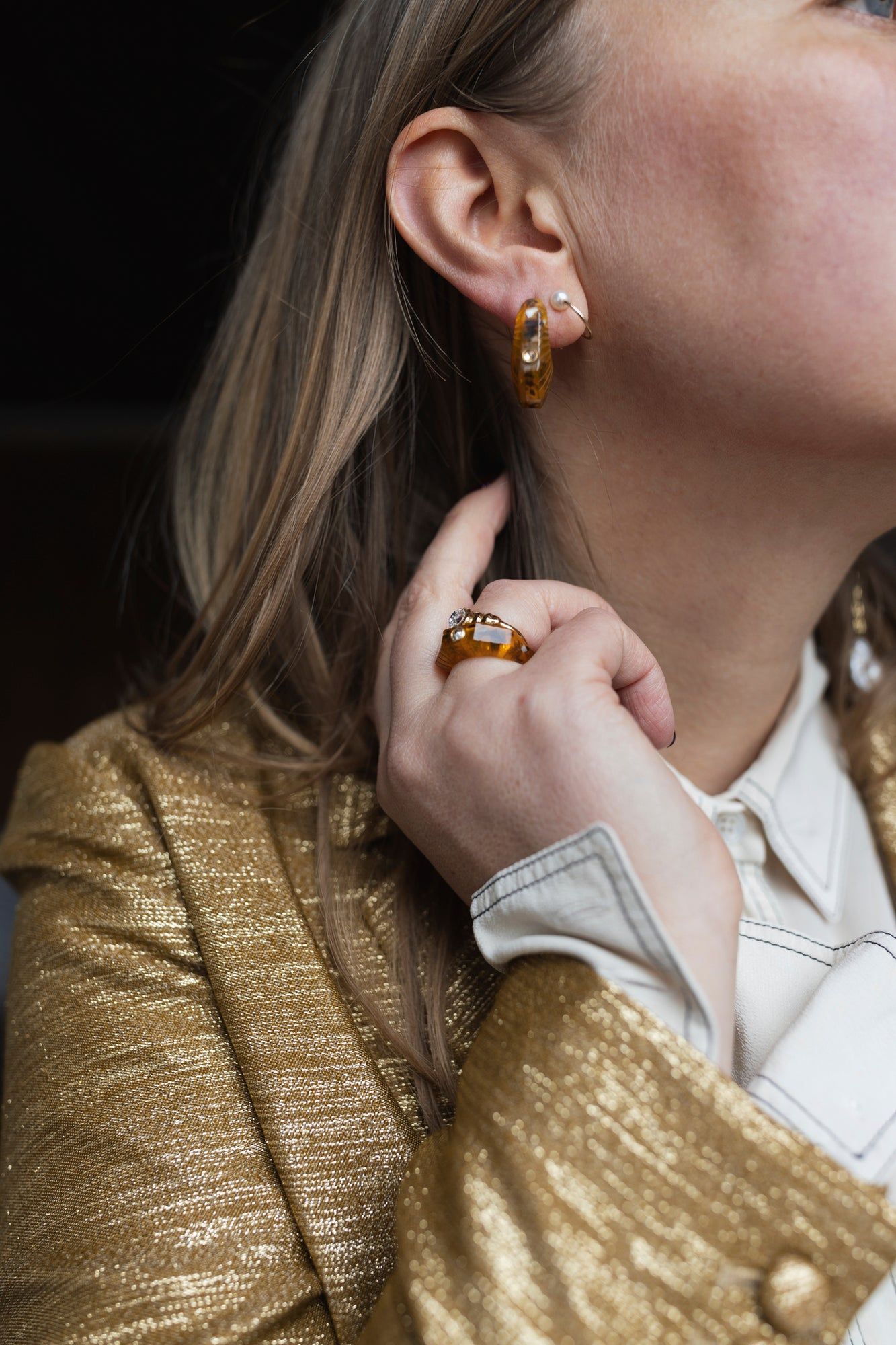 A person wearing a gold textured blazer, white shirt, and WALD Berlin's Isla Bonita Shining Star earrings complements their look with a gold ring adorned with stones in luminous amber while gently touching their ear.
