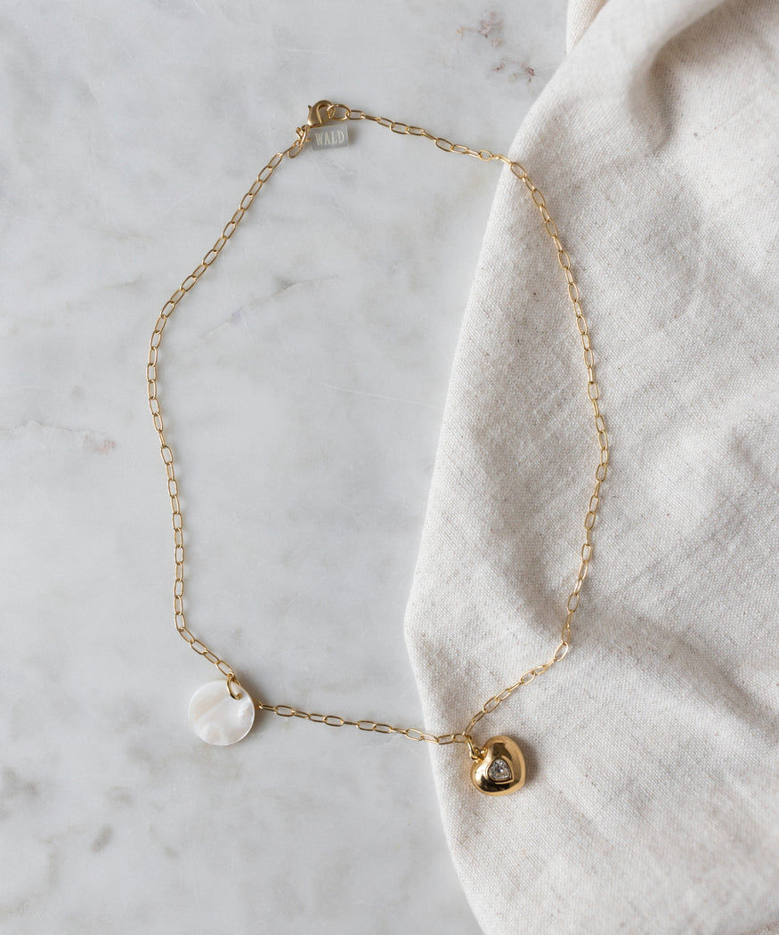 The WALD Berlin Hotel Amour Heart Medallion Necklace features a gold chain with a round mother of pearl pendant and a small heart charm, shown on light marble beside beige fabric.