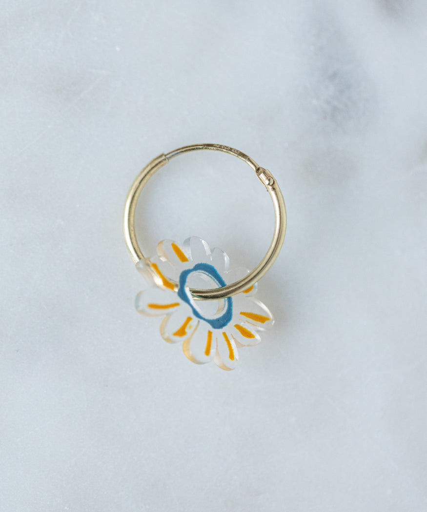 A small gold hoop earring with a translucent flower charm adorned with white petals and yellow accents, crafted from recycled gold, lying on a light gray surface. This product is the Daisy Earrings Transparent by WALD Berlin.