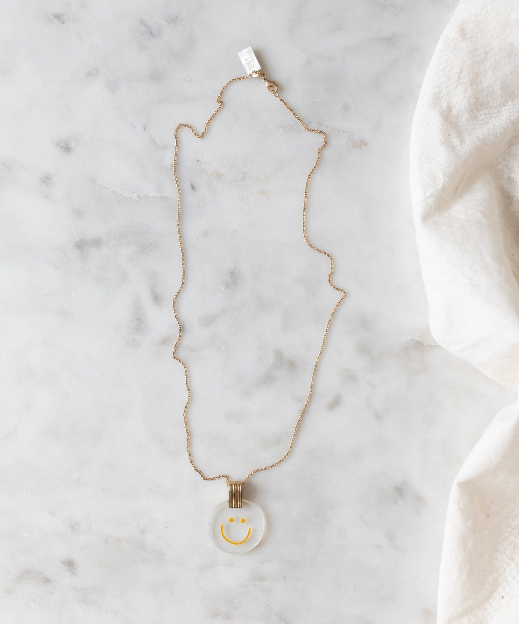 The PRE ORDER Smilie Dude Medaillon Gold Necklace by WALD Berlin, featuring a round pendant with a smiling face, is laid out on a light-colored marble surface, expertly crafted and made in Germany.