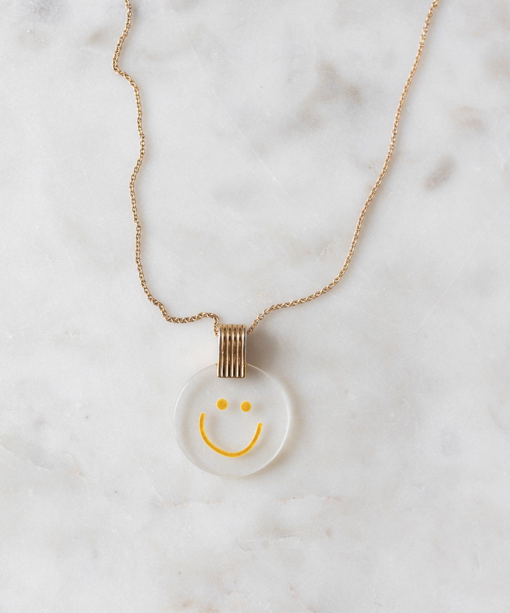 The PRE ORDER Smilie Dude Medaillon Gold Necklace by WALD Berlin features an 18k gold-plated chain with a round transparent pendant displaying a cheerful yellow smiley face, set against a white marble background.