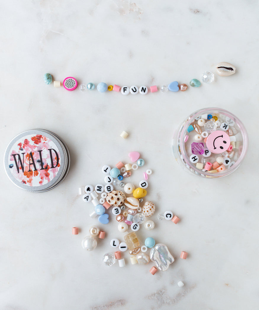 Assorted beads and a bracelet spelling out 'FUN' are arranged on a marble surface. Nearby, there is a tin labeled 