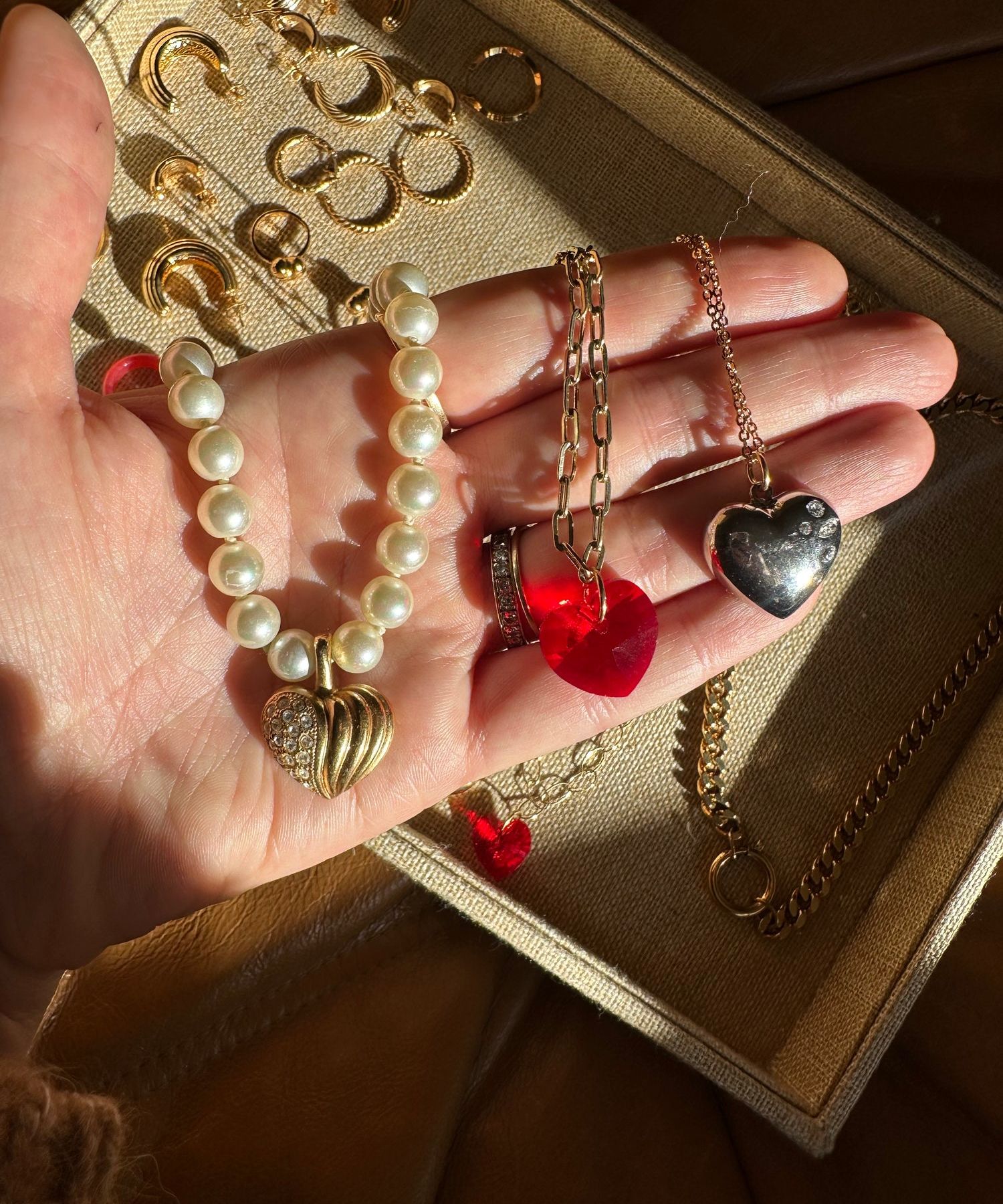 A hand displays a pearl bracelet with gold charms, earrings, a red heart pendant, and a silver ring. More jewelry rests in a beige tray beside the chic surprise WALD Berlin Bag by WALD Berlin.