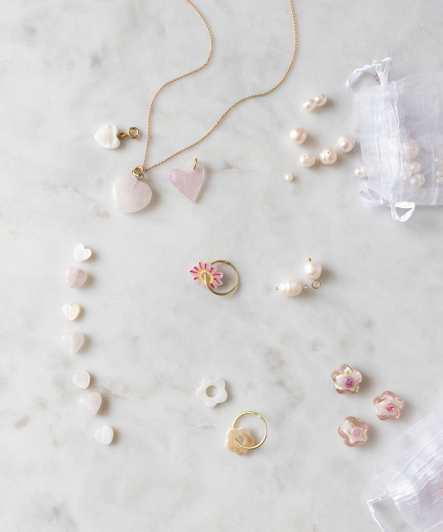 Assorted jewelry, including heart-shaped pendants, rings, pearl earrings, and the WALD World Mother of Pearl Heart Beads Set, are spread out on a marble surface. A small sheer bag contains additional WALD World mother of pearl necklaces and other jewelry items.