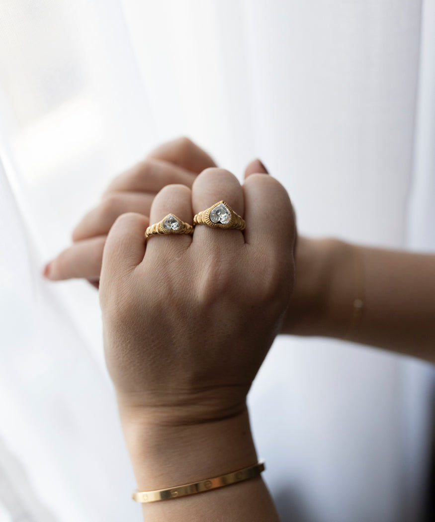 A person's hand with two WALD Berlin Be My Lover Gold Rings, one on the ring finger and one on the middle finger, against a white sheer curtain.