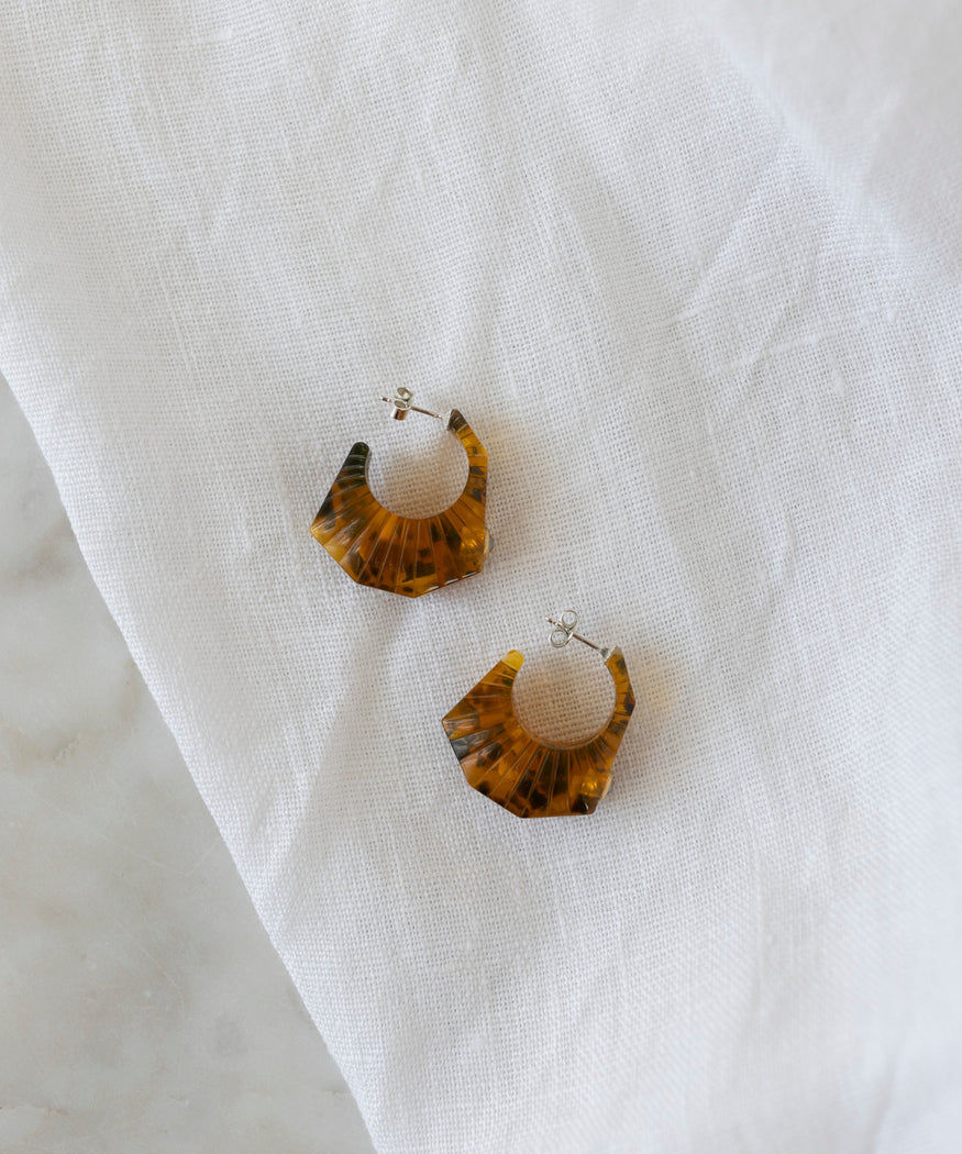 The Isla Bonita Shining Star earrings by WALD Berlin, crafted from bio polyester, showcase two brown, semi-transparent hoops with a geometric design resting elegantly against a white fabric backdrop.