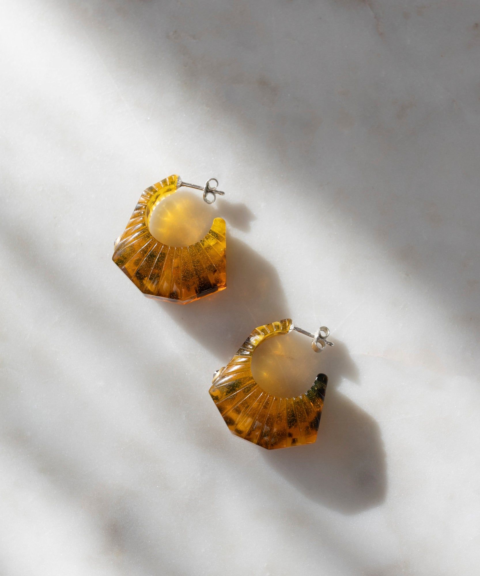 A pair of amber, fan-shaped Isla Bonita Shining Star earrings from WALD Berlin is placed on a light marble surface, illuminated by soft sunlight.