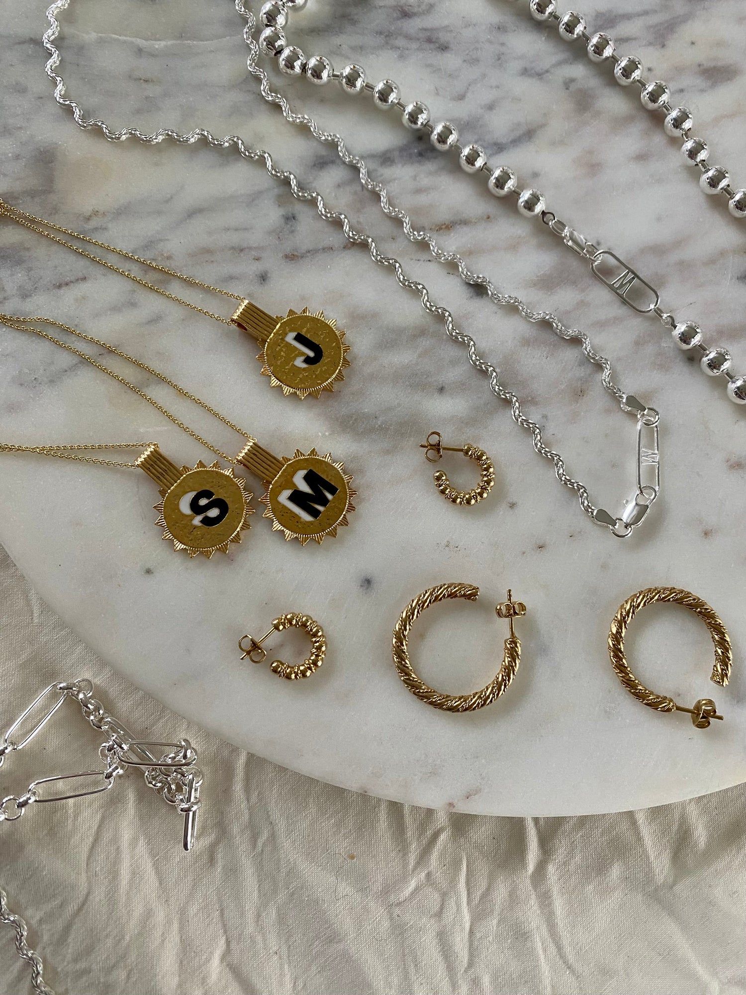 The Naomi Bracelet Silver by WALD Berlin is displayed on a marble surface alongside initial necklaces, textured hoop earrings, and various recycled gold and silver chain styles.