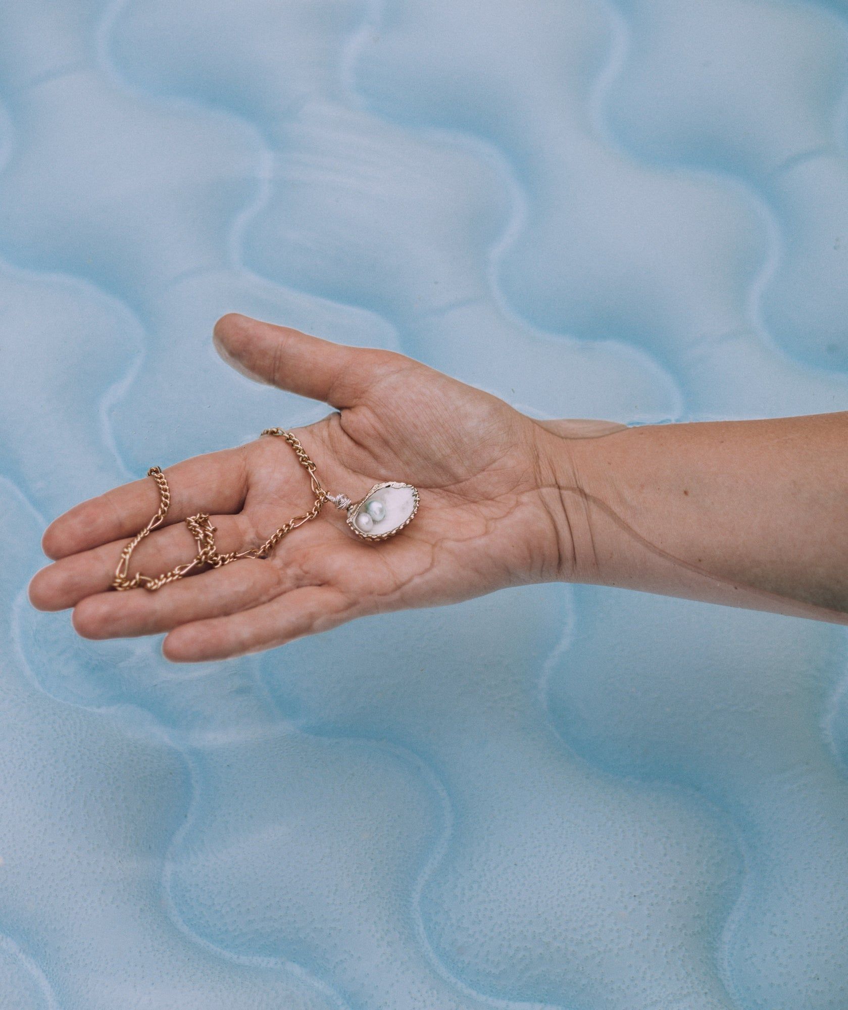 A hand holding a delicate Drop It Like It’s Hot Shell Necklace by WALD Berlin with a pendant over a blue water background.