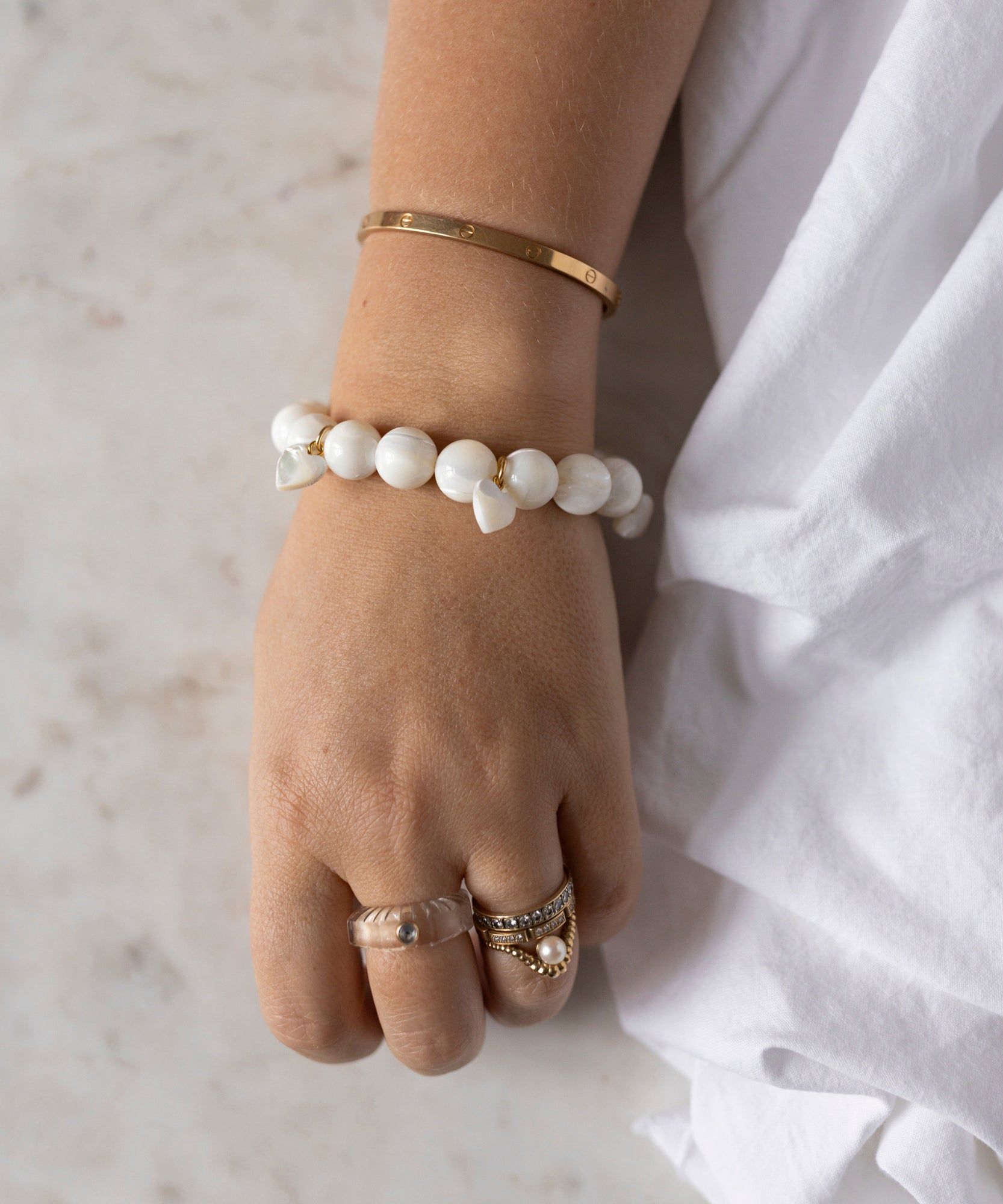 A hand wearing a WALD Berlin Mother Of Pearl Multi Heart Bracelet, a gold bangle, and three rings rests on a light surface. The person is dressed in white fabric.