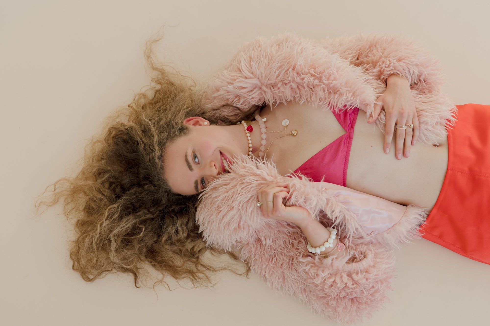 A person with curly hair relaxes on a cream surface, wearing a fluffy pink jacket, pink top, and red pants. They accessorize with the WALD Berlin Mother Of Pearl Multi Heart Bracelet and other jewelry.