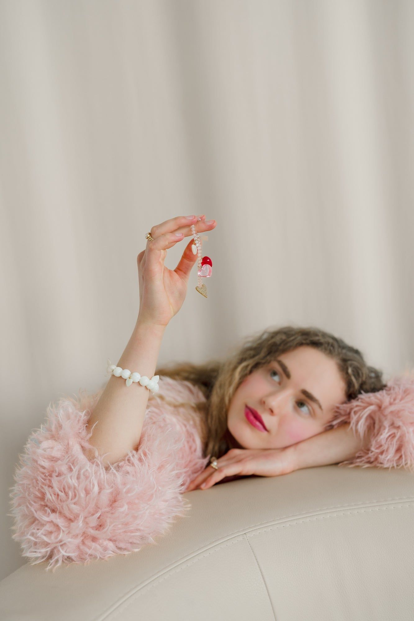 A person rests their head on their arm while lying on a couch, wearing a fluffy pink sweater and holding the WALD Berlin Mother Of Pearl Multi Heart Bracelet, featuring a small red and white design with a gold heart charm.