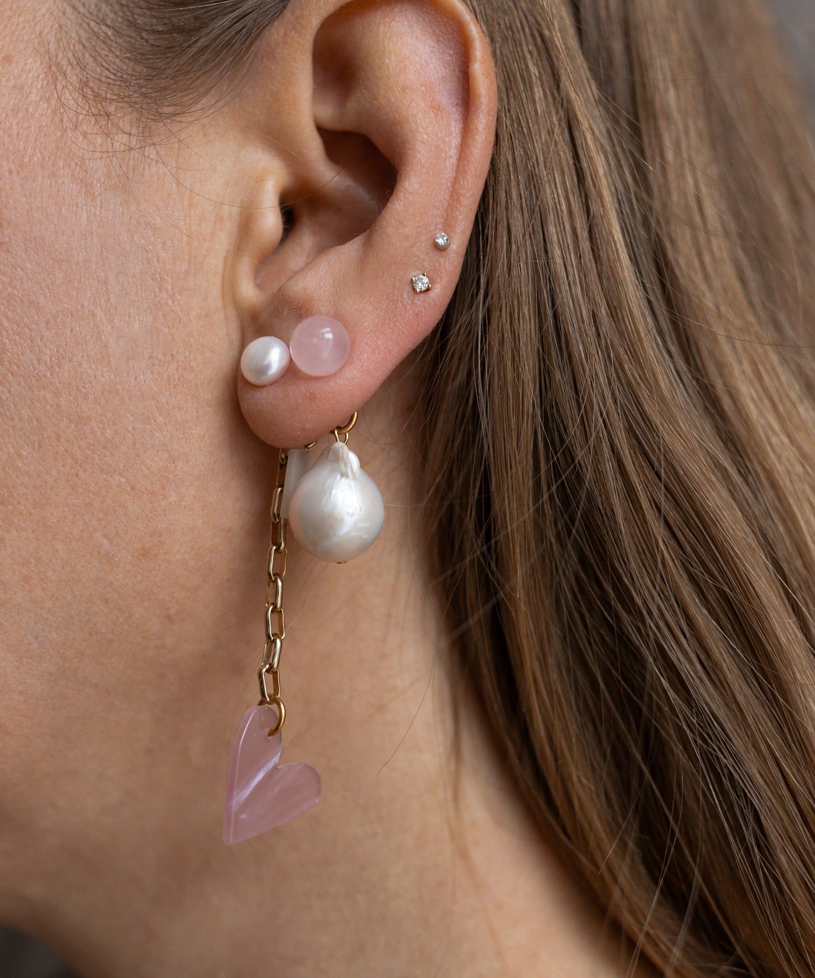 A close-up of a person's ear displays the XL Pearl Heart I Am Love Earrings by WALD Berlin, featuring multiple piercings adorned with an XL sweet water pearl, studs, a dangling charm, chain, and pink heart. Artfully crafted and proudly made in Germany.
