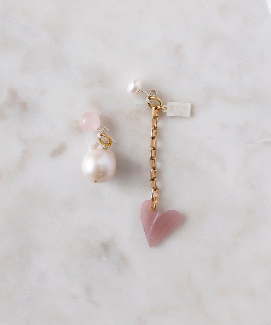 On a marble surface, WALD Berlin's XL Pearl Heart I Am Love Earrings feature one with a gold chain and pink heart pendant, and another showcasing an XL sweet water pearl with mixed pink and white pearls, exemplifying stylish craftsmanship made in Germany.