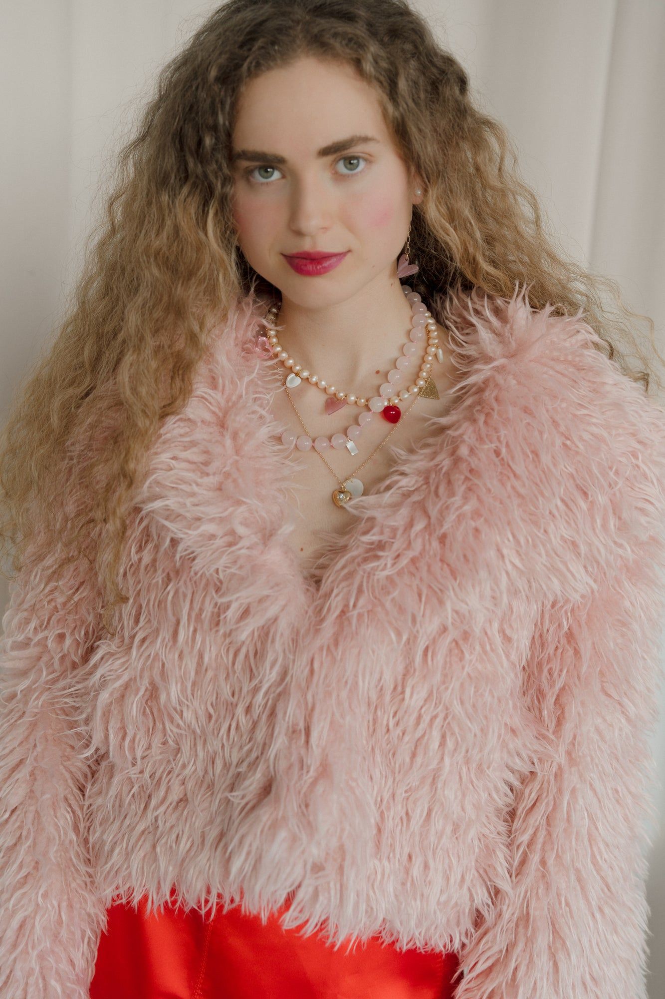 With long curly hair, a person dons a fluffy pink jacket and layered necklaces, highlighted by WALD Berlin's XL Pearl Heart I Am Love Earrings, set against a plain background.