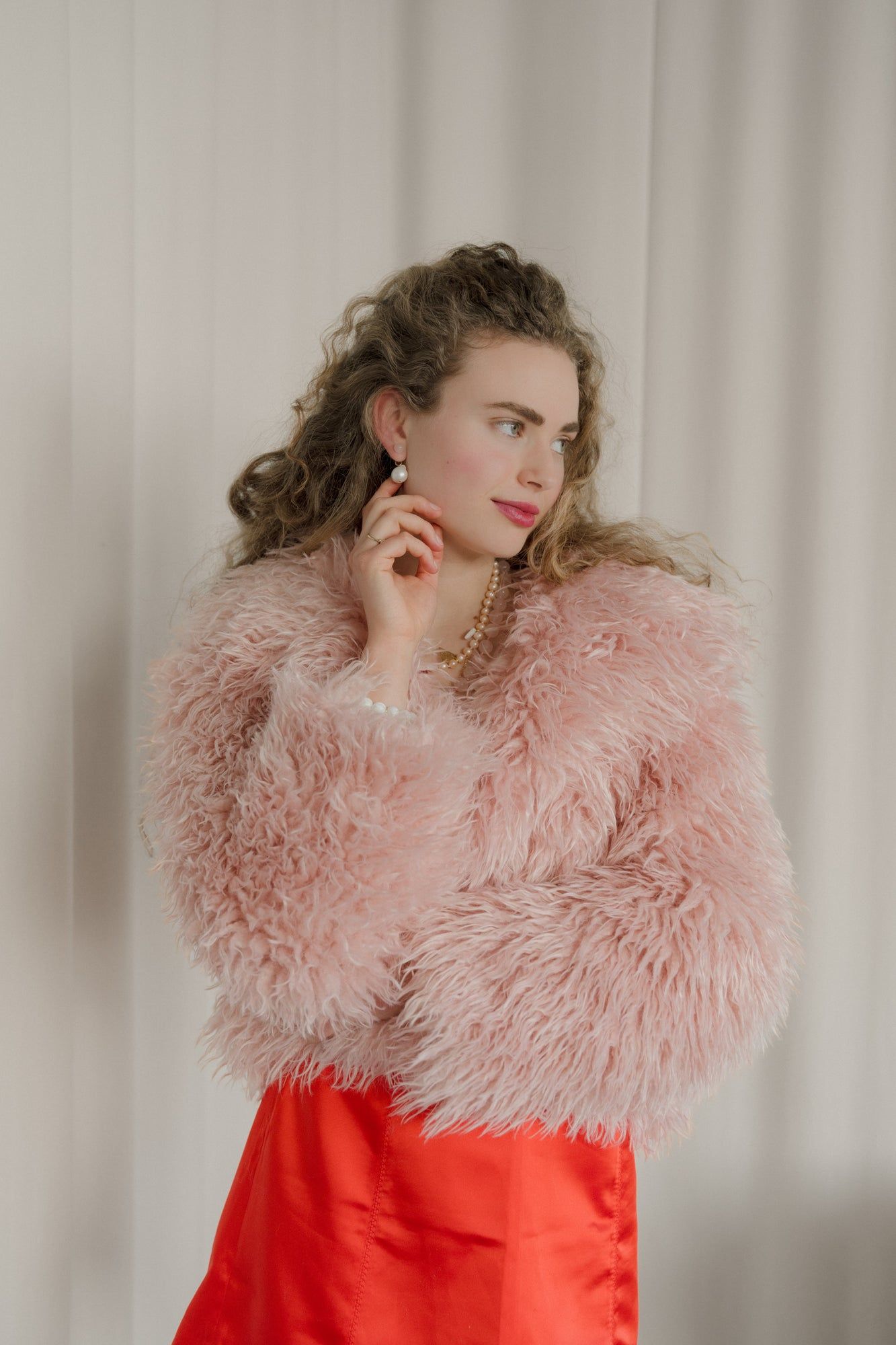 A person poses against a light background, wearing a fluffy pink jacket and red dress, beautifully highlighted by XL Pearl Heart I Am Love Earrings from WALD Berlin.
