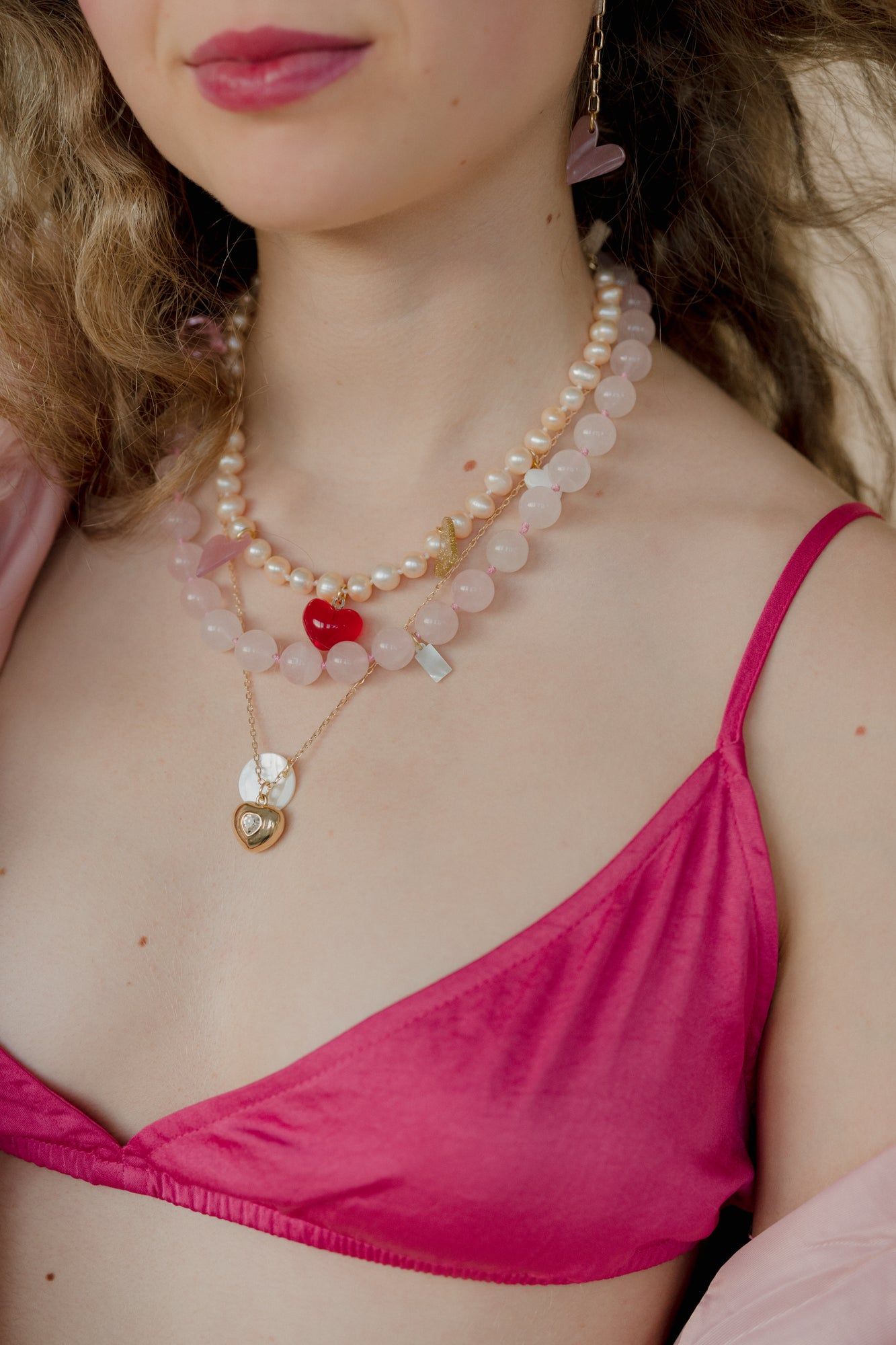 A person wearing a pink top, two heart-shaped necklaces, and WALD Berlin's XL Pearl Heart I Am Love Earrings with an XL sweet water pearl. Their curly hair complements their slightly visible smile.