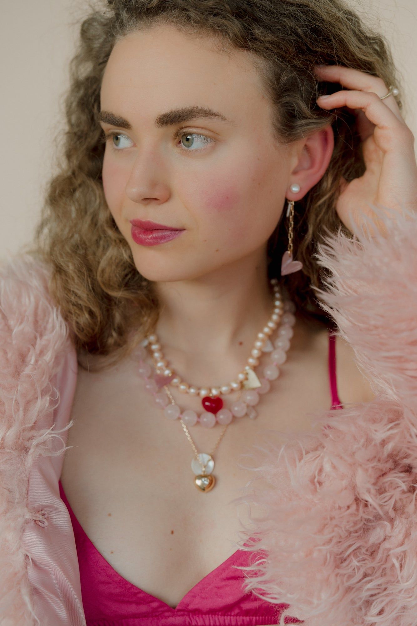 A woman in a pink fur coat with pearl heart pendant jewelry glances sideways, elegantly showcasing an XL sweet water pearl from the mix-and-match WALD Berlin 