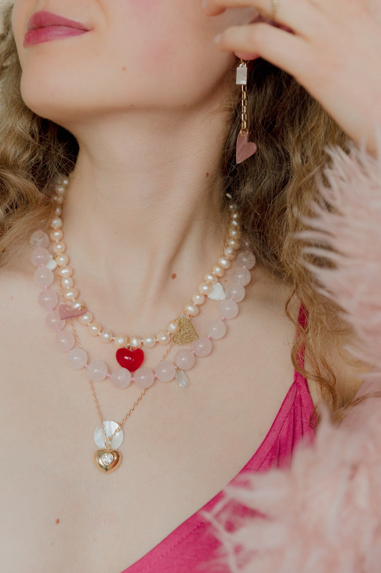 A woman wearing layered necklaces with XL sweet water pearls and heart-shaped pendants, a pink top, and a fluffy pink garment effortlessly showcases her mix and match style, including WALD Berlin's XL Pearl Heart I Am Love Earrings.