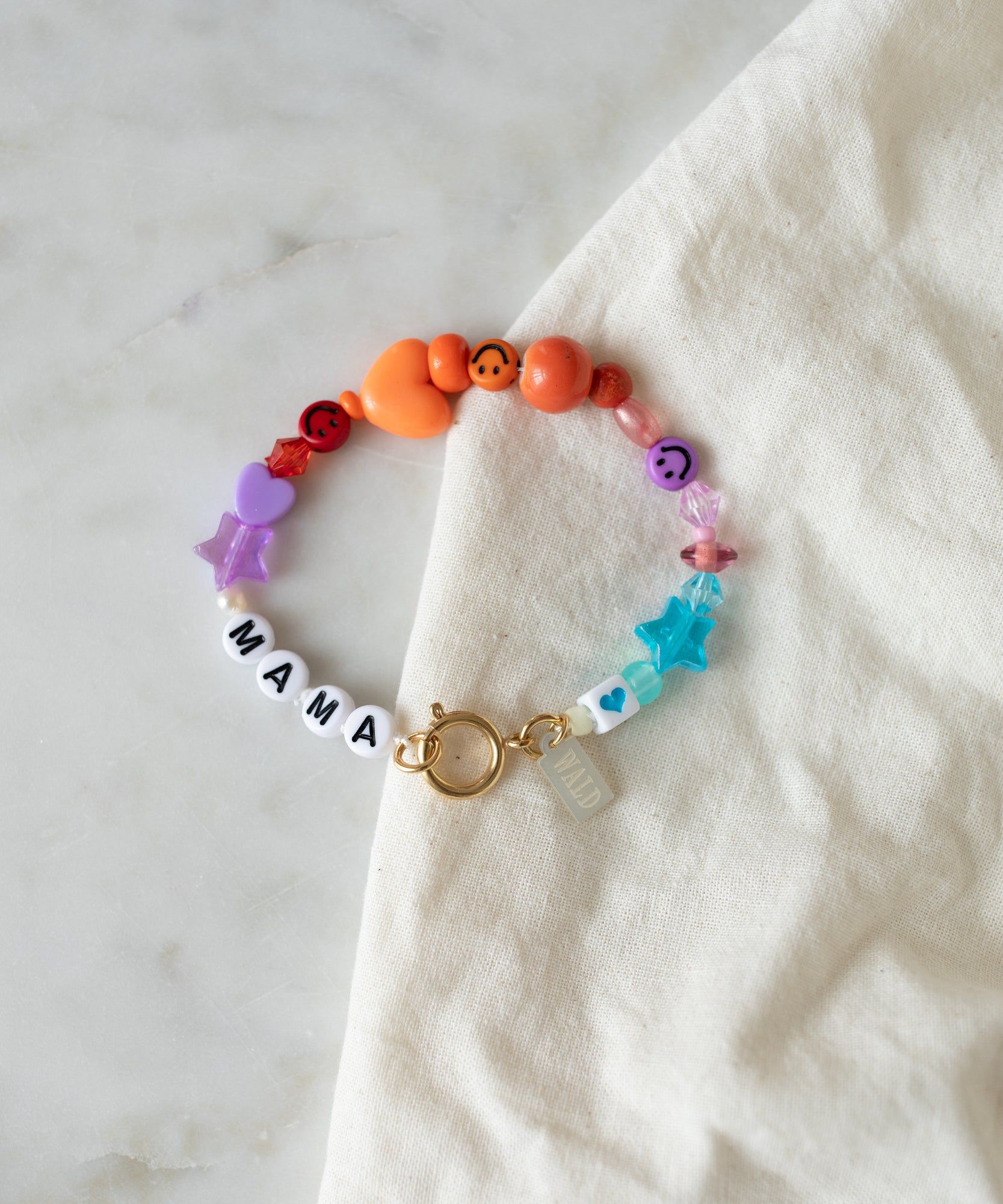 A handmade, colorful Limited Mama Bracelet by WALD Berlin adorned with various charms, laid on a cream fabric.