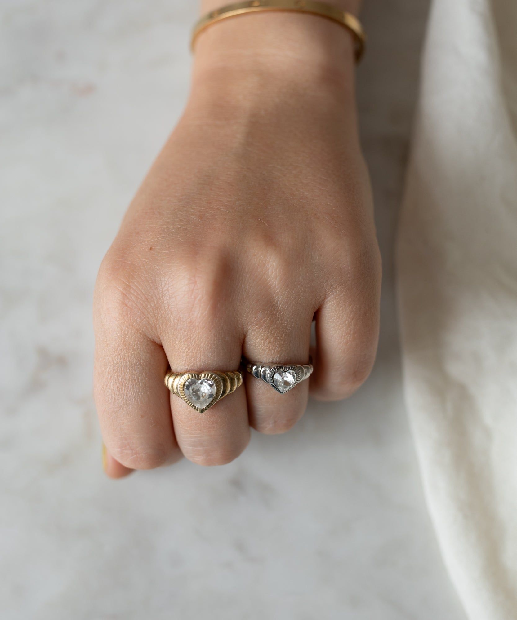 A close-up image of a hand wearing two detailed rings, one Be My Lover Mini Silver Ring by WALD Berlin and one gold, resting on a marble surface.