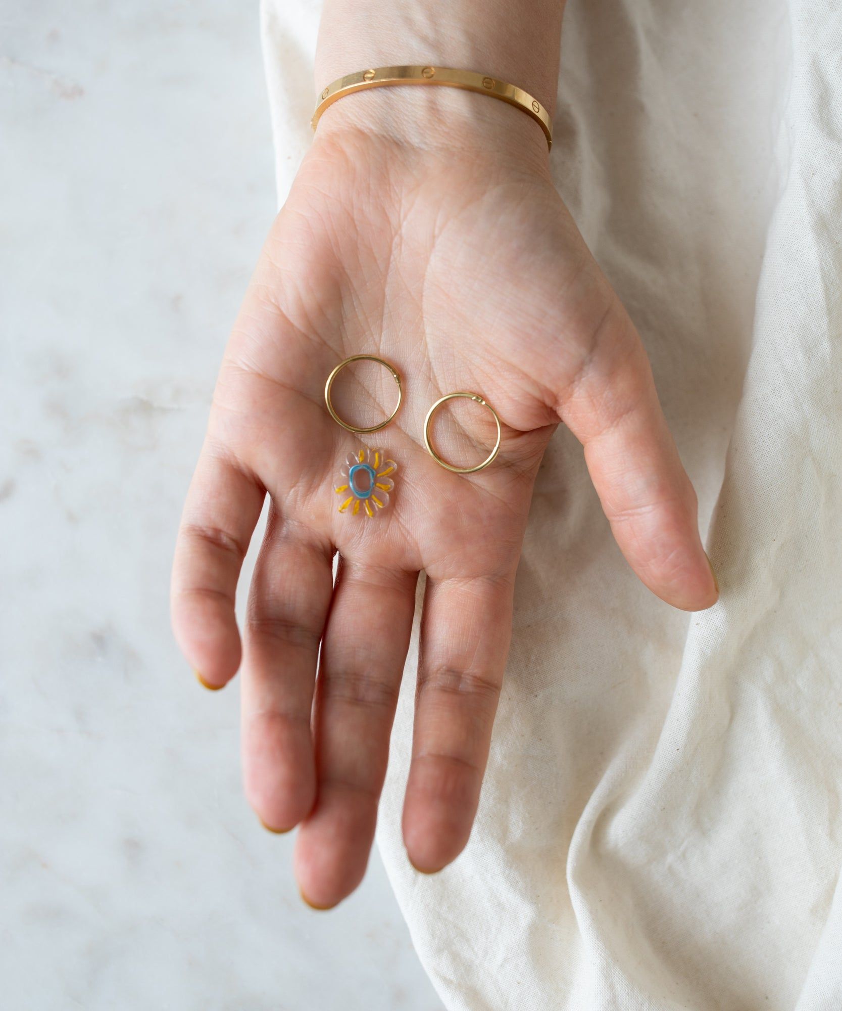 A person's open hand displaying three rings: two gold bands and one with a blue gem, against a cream fabric backdrop featuring WALD World's Daisy Transparent Charm.