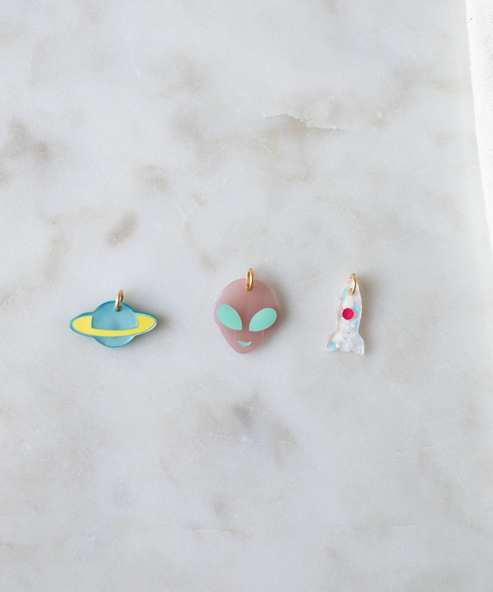 Three quirky Rocket Charms placed on a marble surface: one shaped like a planet, another as an alien head, and a third resembling a clear ghost with a red eye from the WALD World collection.