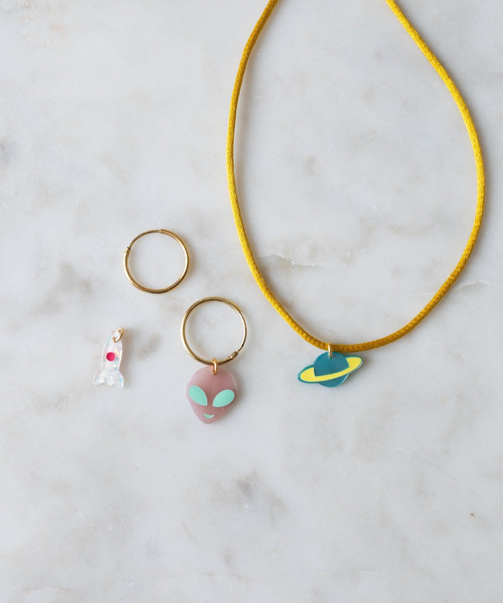 Two Venus Charm gold rings with colorful pendants and a yellow necklace from WALD World on a marble surface.