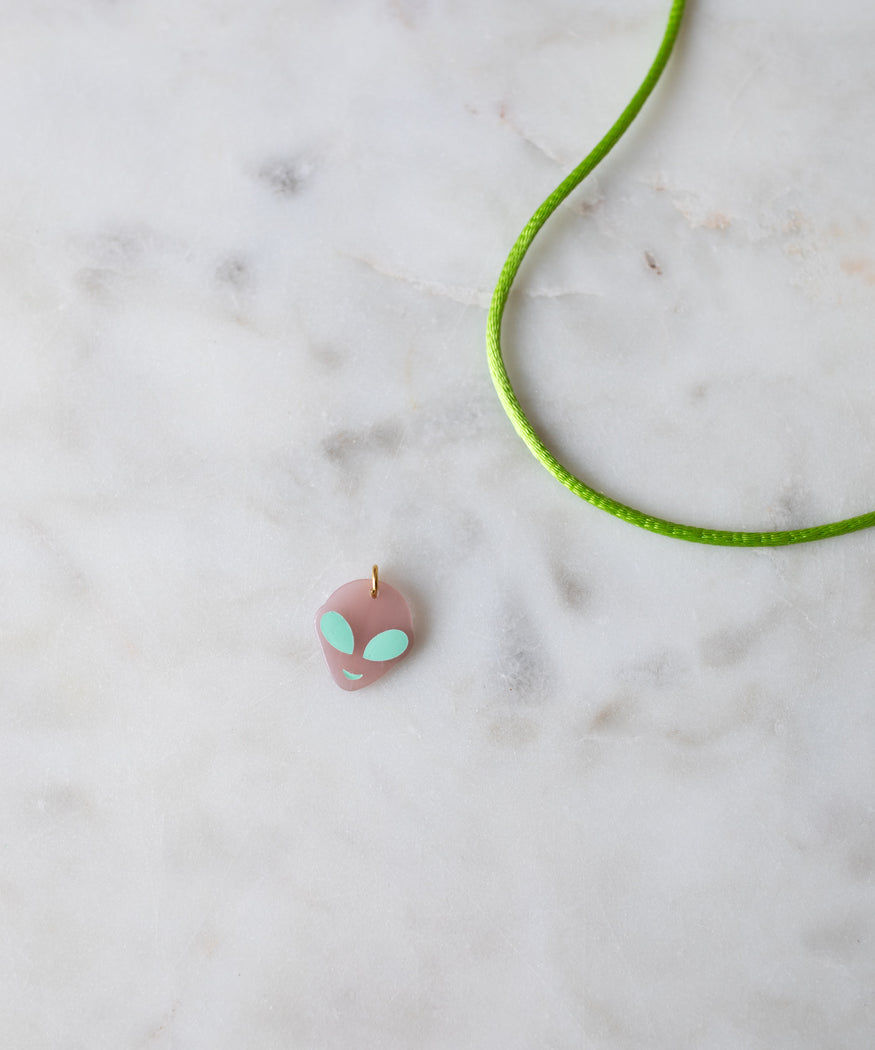 A pink, Alien Charm pendant on a green cord, crafted as DIY jewelry by WALD World, lying on a marble surface.