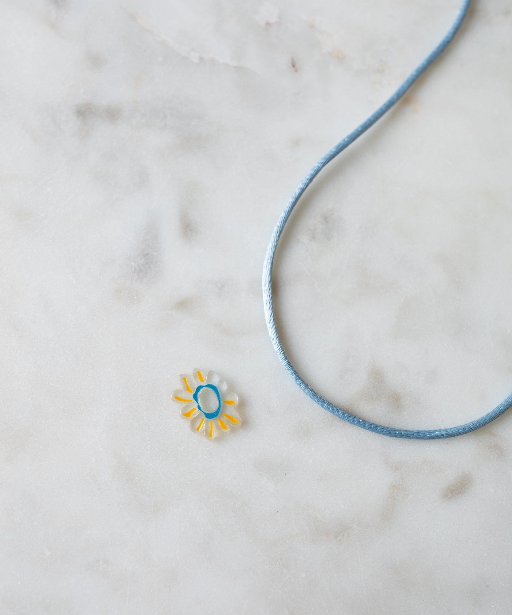 A Daisy Transparent Charm pendant with a turquoise center and a blue cord on a marble surface, ideal for DIY jewelry enthusiasts by WALD World.