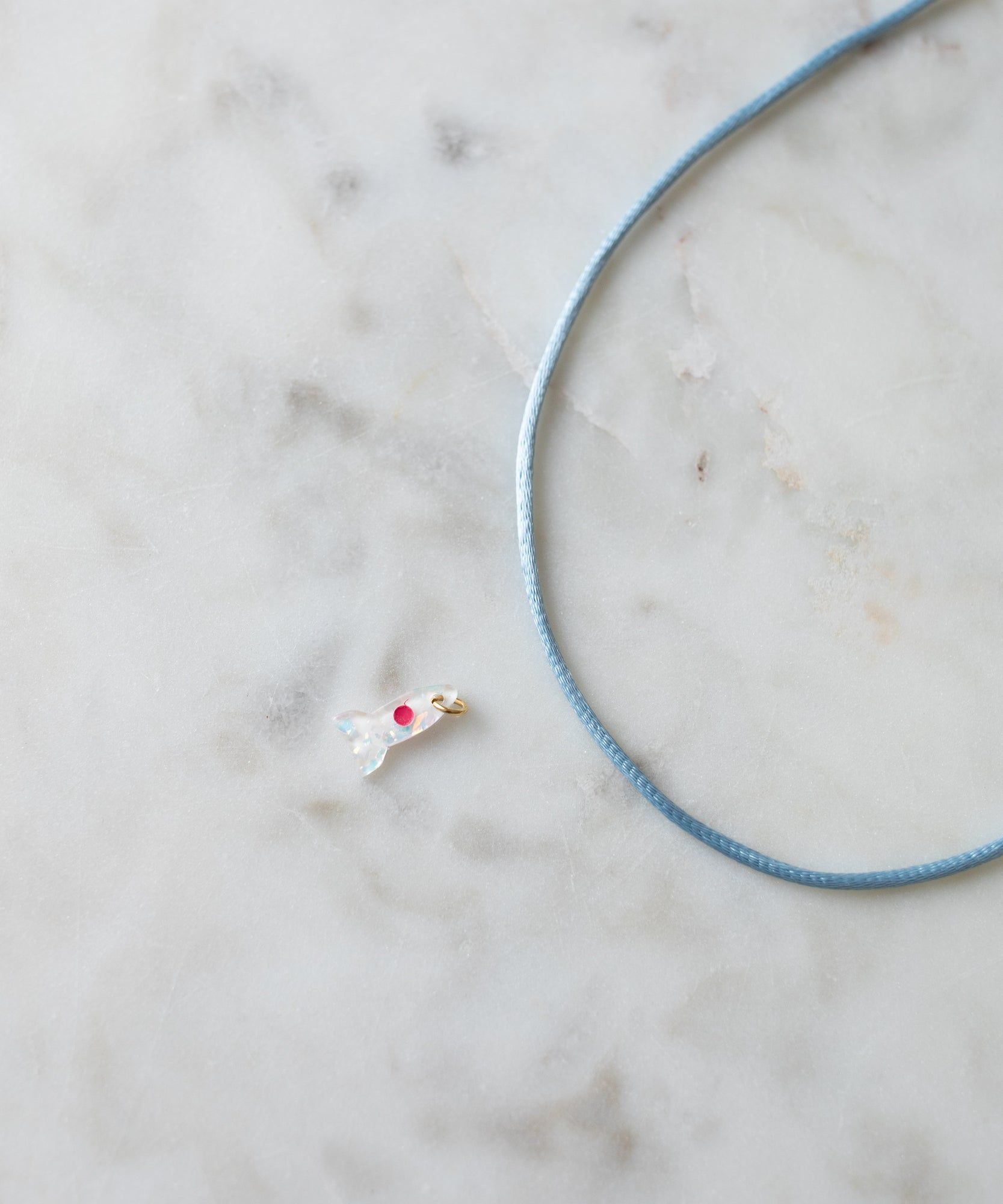 A delicate Rocket Charm pendant with a red gem, shaped like a bird, lies next to a light blue cord on a marbled surface, epitomizing the essence of DIY jewelry from WALD World.