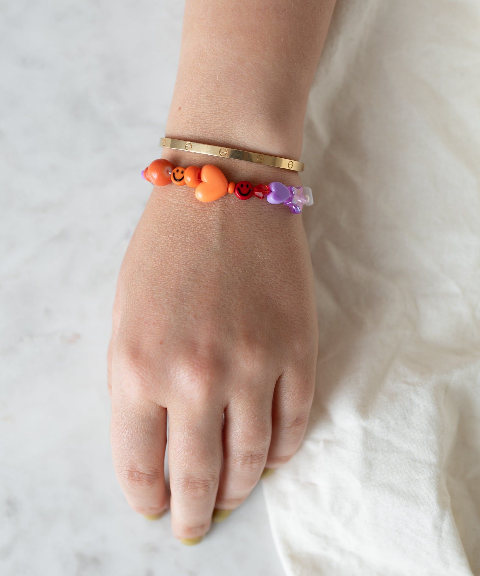 A person's wrist adorned with a WALD Berlin Limited Mama Bracelet featuring orange and purple charms, resting on a white textured surface.