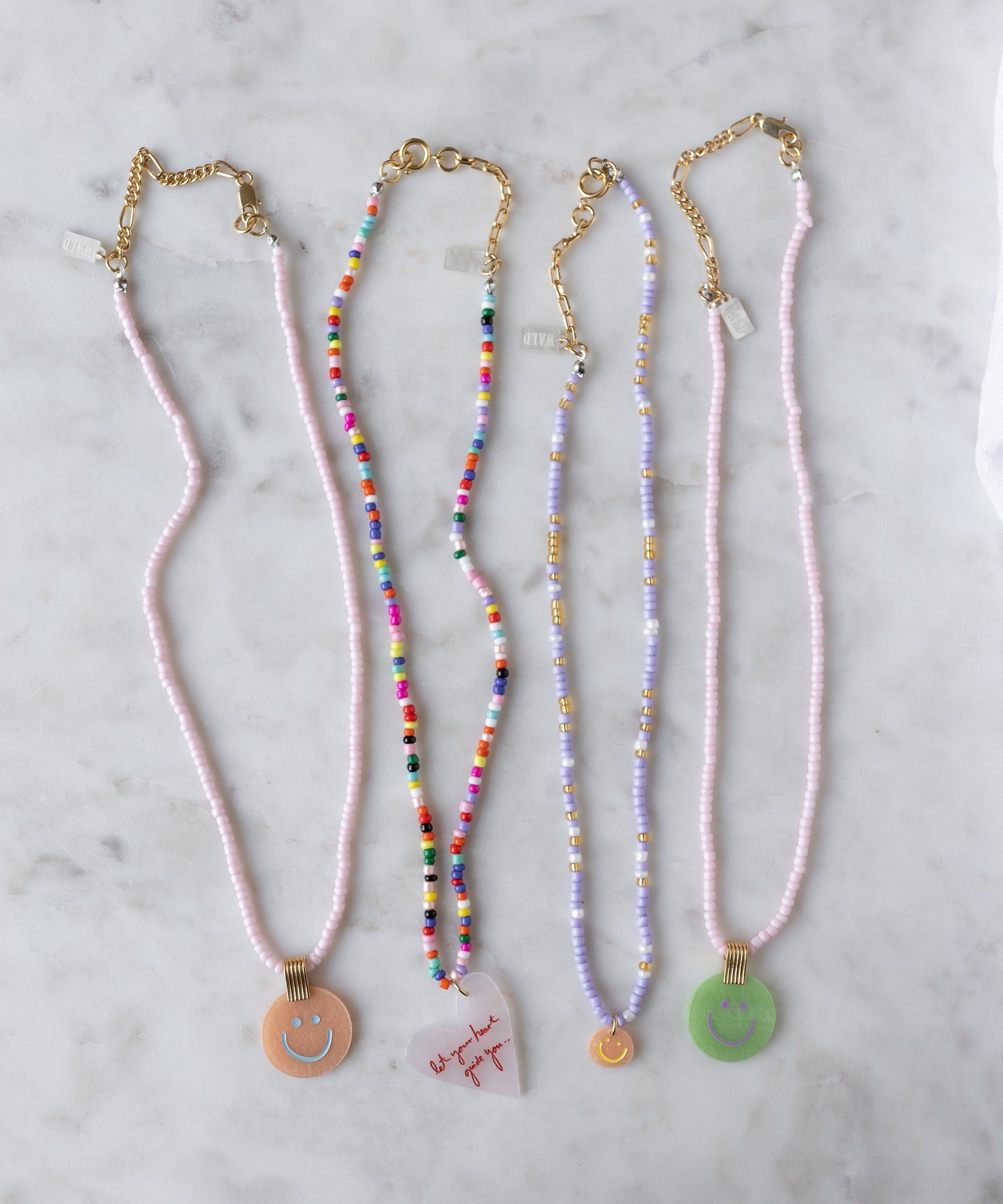 Four WALD Berlin necklaces with gold clasps rest on marble: three feature the Pop Smilie Medallion Peach pendant, while one is adorned with a heart and red text for a playful summer touch.