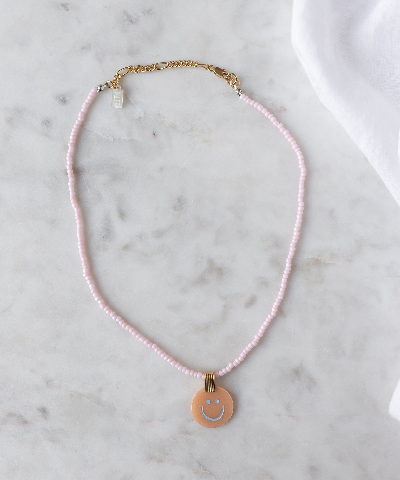 The Pop Smilie Medallion Peach by WALD Berlin is a pink beaded necklace with a gold clasp and round pendant featuring a blue smiley face—ideal for adding playful charm to classic styles or your summer look.