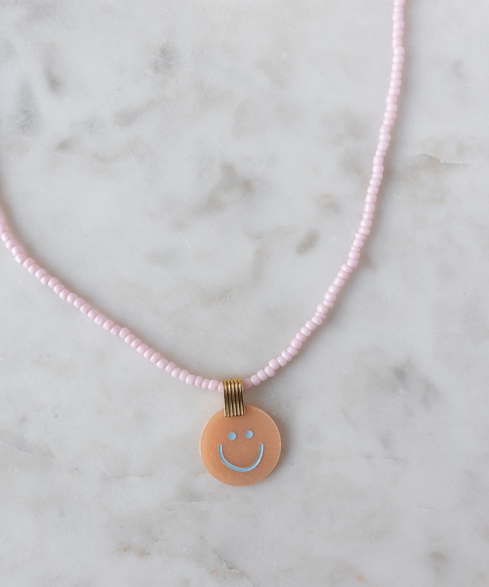 Add a cheerful pop to your style with the Pop Smilie Medallion Peach necklace from WALD Berlin—a pink beaded piece with a round peach pendant, blue smiley face, and playful vibes, perfect for brightening up your summer look.