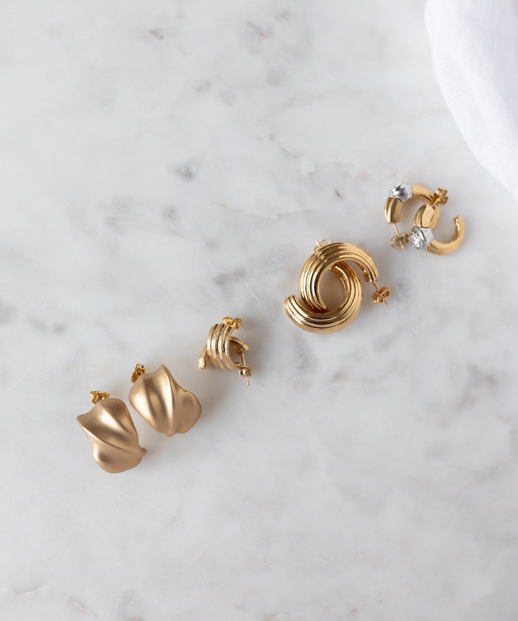 Four pairs of WALD Berlin earrings, including the Pop Wave Earrings Small in gold and abstract shapes with 925 silver pin posts, are arranged on a white marble surface.