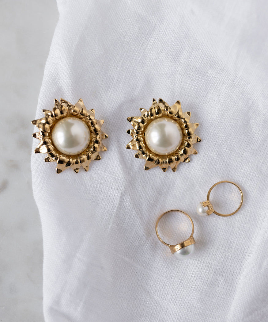The Pop Soleil Earrings by WALD Berlin, featuring large pearl centers and spiked gold edges, are displayed alongside two 18k gold plated rings with single pearls on a white fabric background.
