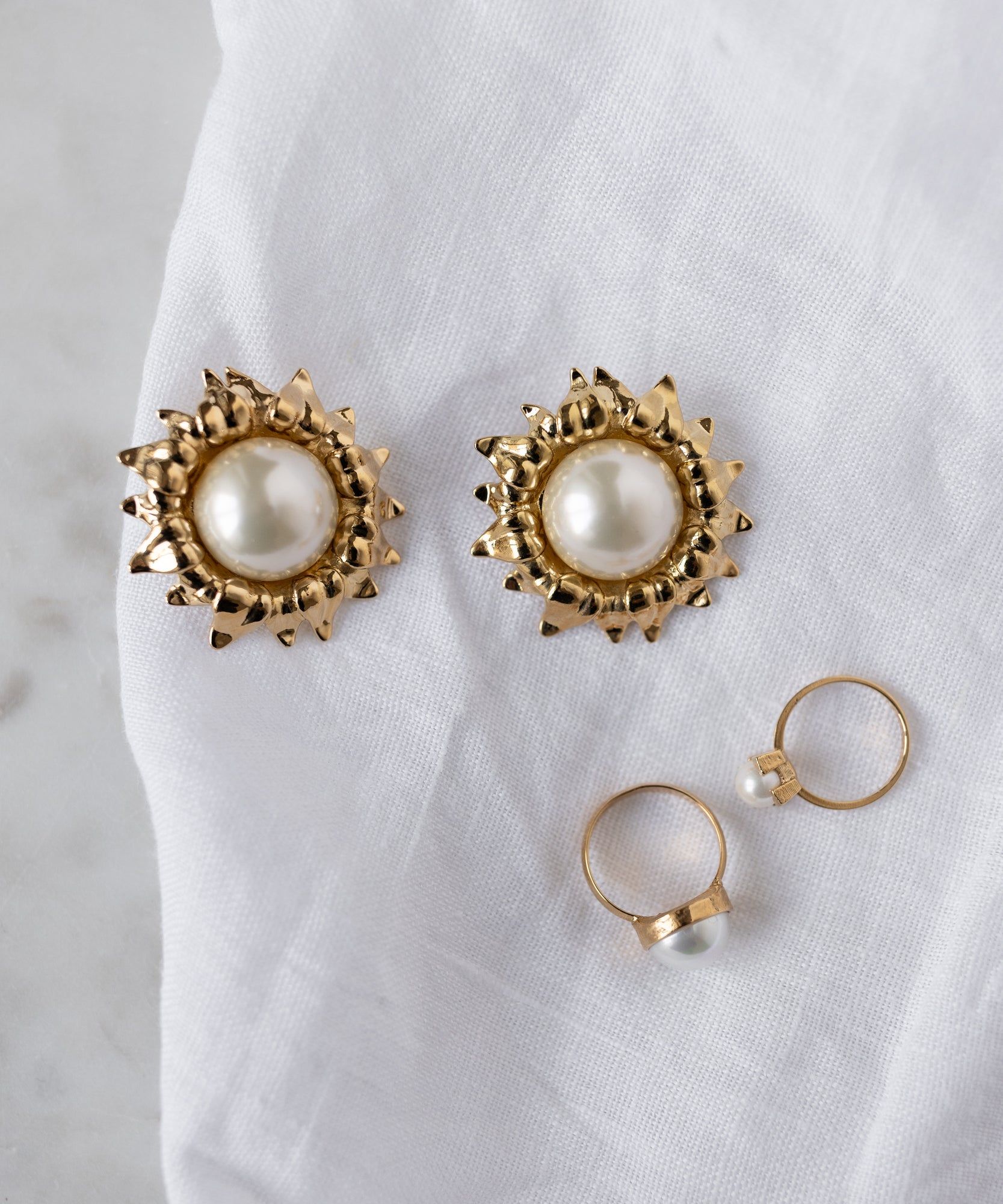WALD Berlin Pop Pearl Ring Mini: Two 18k gold plated brass rings with small pearl accents, paired with gold earrings featuring large pearl centers, displayed on a white fabric background.