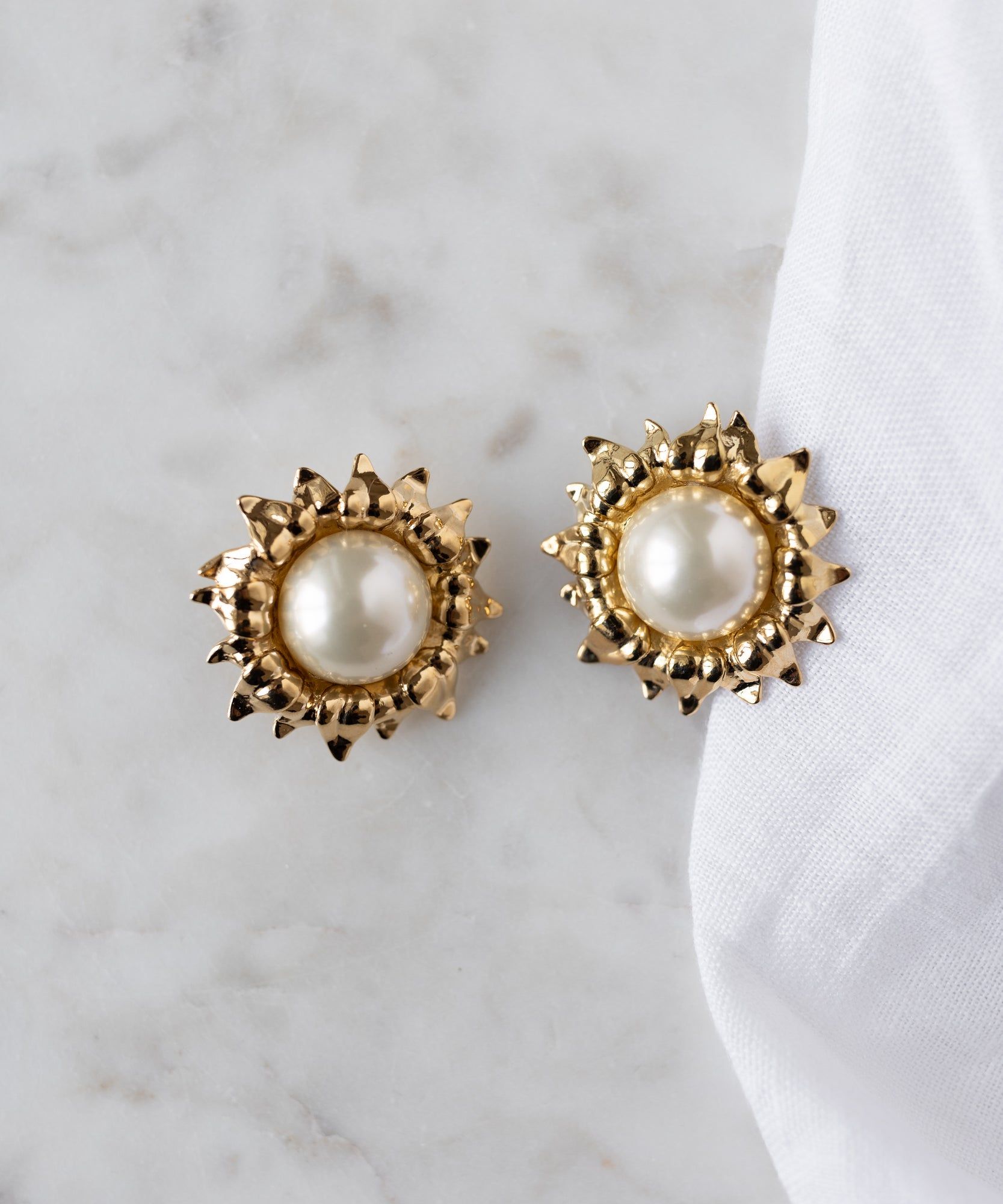 The Pop Soleil Earrings by WALD Berlin, featuring round 18k gold plating and wax pearl centers, are displayed on a marble surface beside a white cloth.