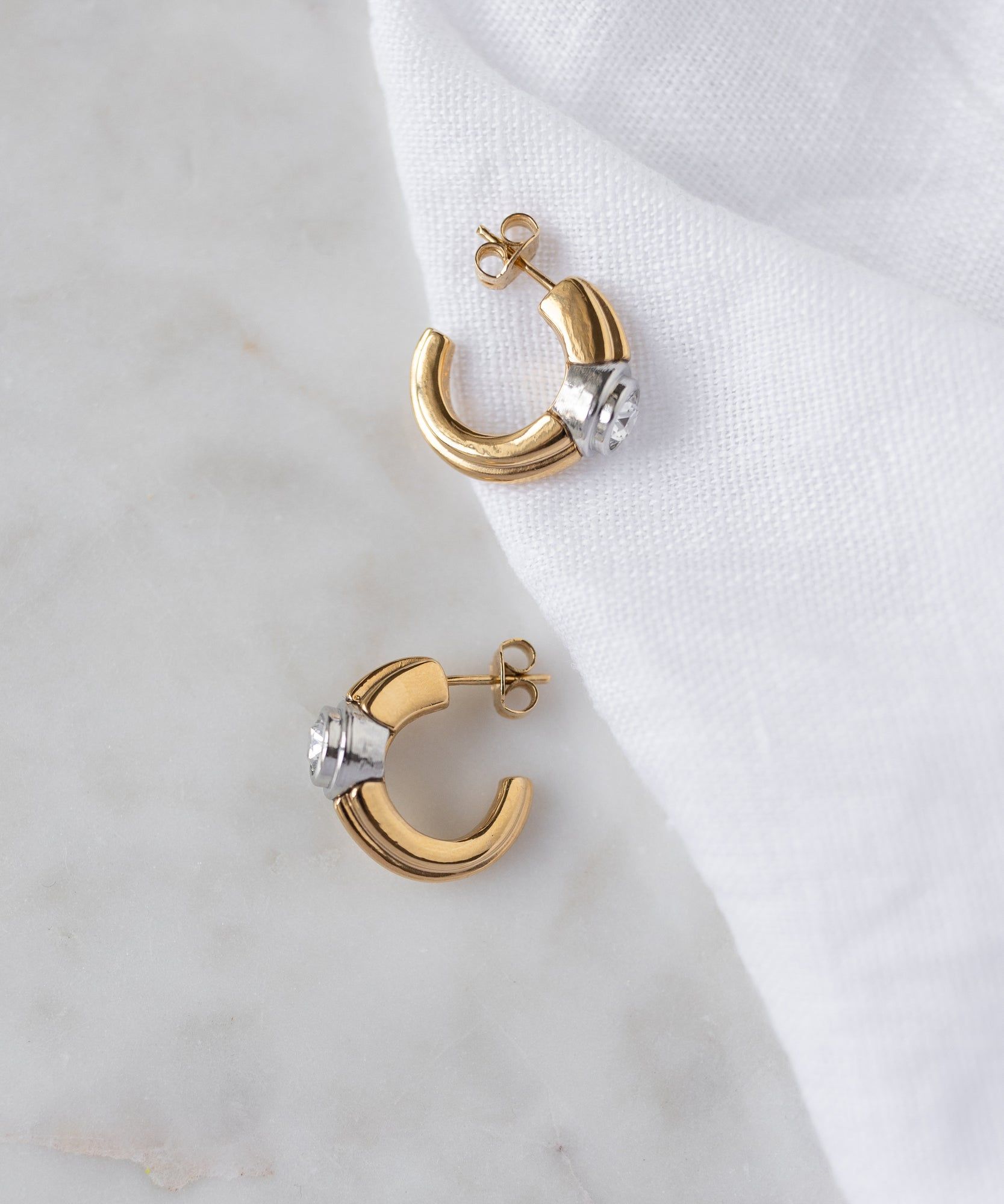 The WALD Berlin Pop Bi Color Crystal Earrings, featuring 18k gold plating and silver hoops with post backings, are displayed on a white fabric and marble surface.