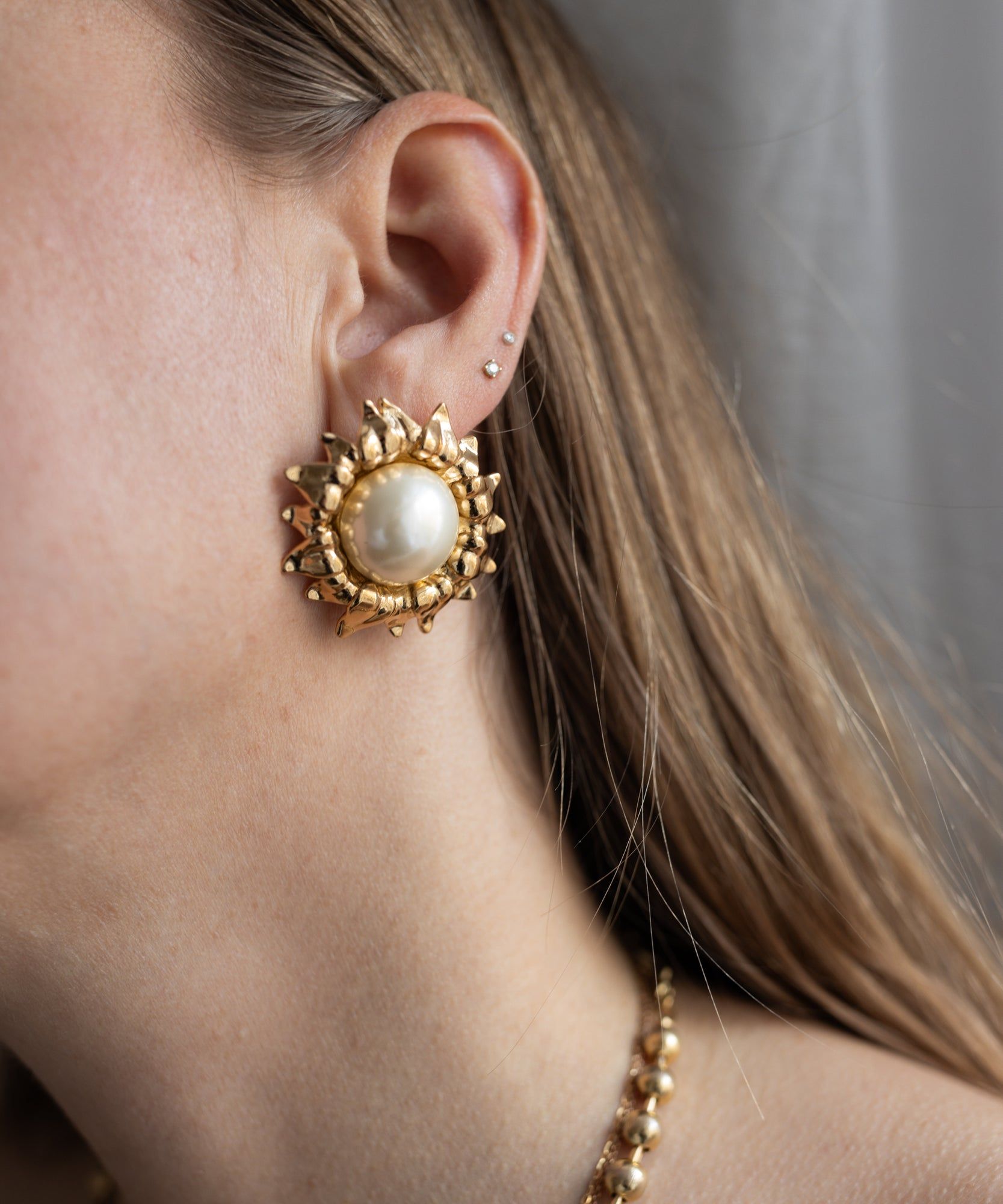 Close-up of a woman's ear wearing WALD Berlin's Pop Soleil Earrings—large, 18k gold plated sunburst design with wax pearl accents. She also wears two small studs in her upper ear and part of a beaded necklace is visible.