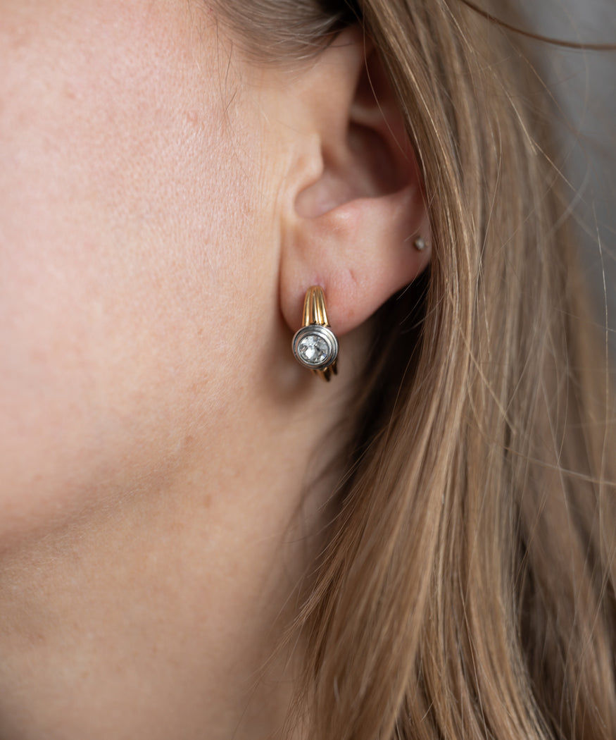 Close-up of a woman's ear wearing WALD Berlin Pop Bi Color Crystal Earrings featuring an 18k gold-plated design with a round, clear Swarovski stone. Partially visible brown hair frames the ear.